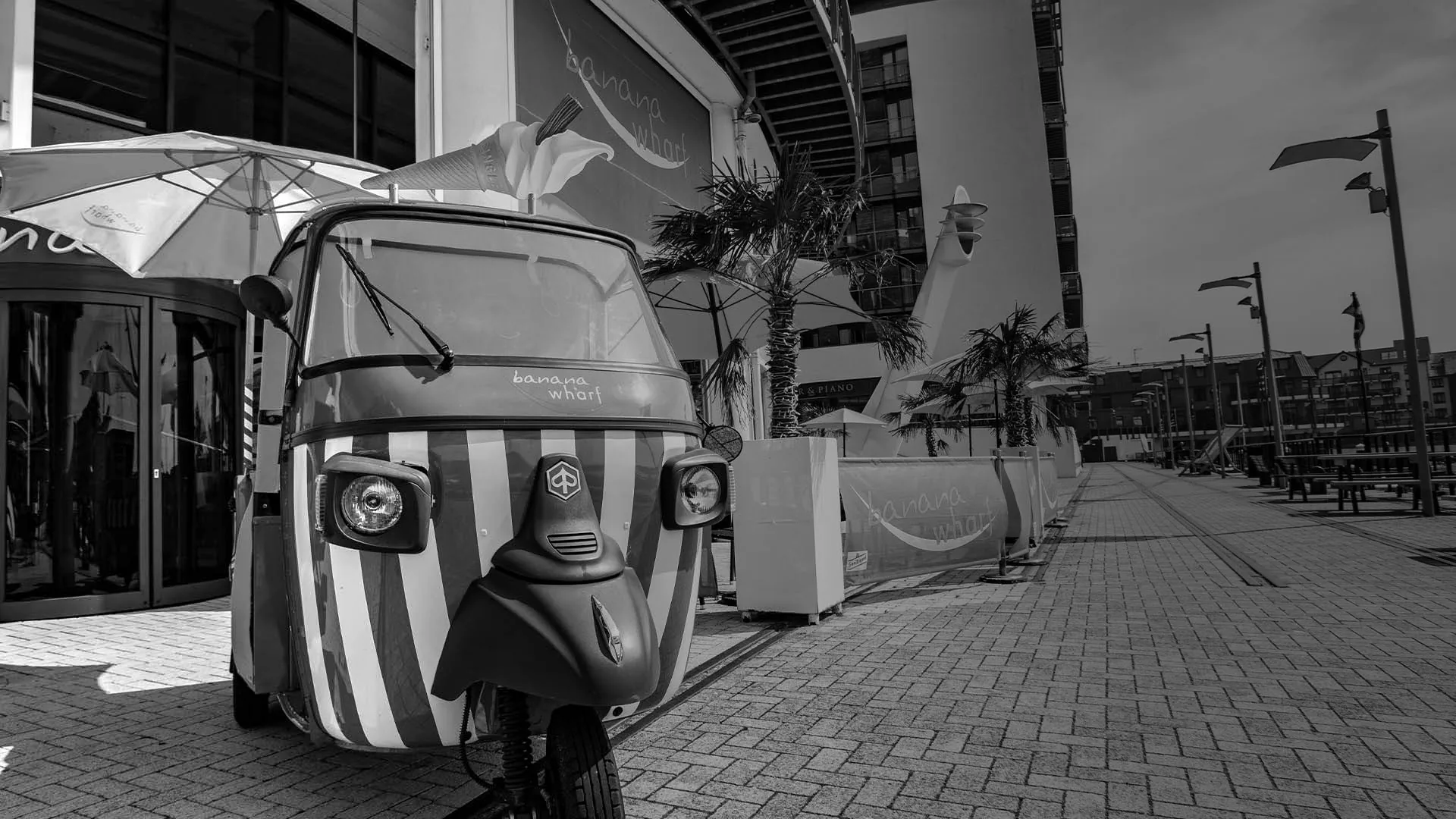 A striped Banana Wharf ice-cream tuk-tuk parked on the Ocean Village boardwalk, palm trees and apartment blocks behind, in black and white
