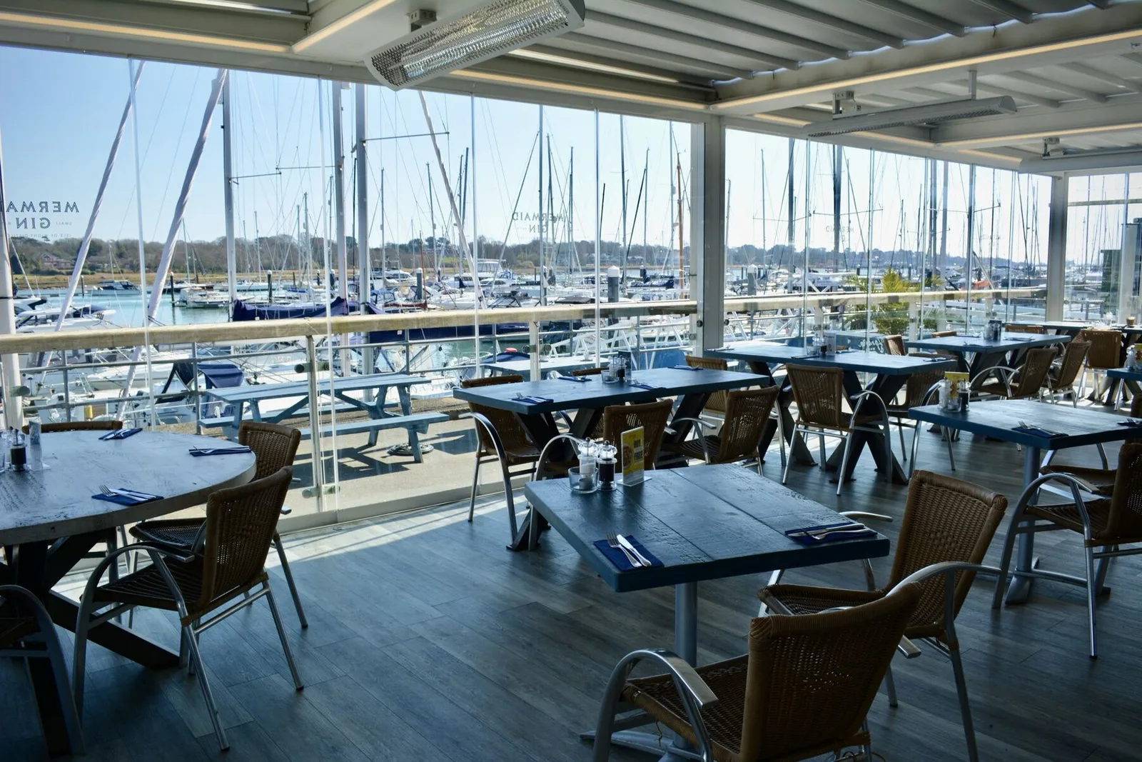 Banana Wharf Hamble's covered terrace on a sunny day, Mermaid Gin branding on the glass sides and boats moored in the marina beyond